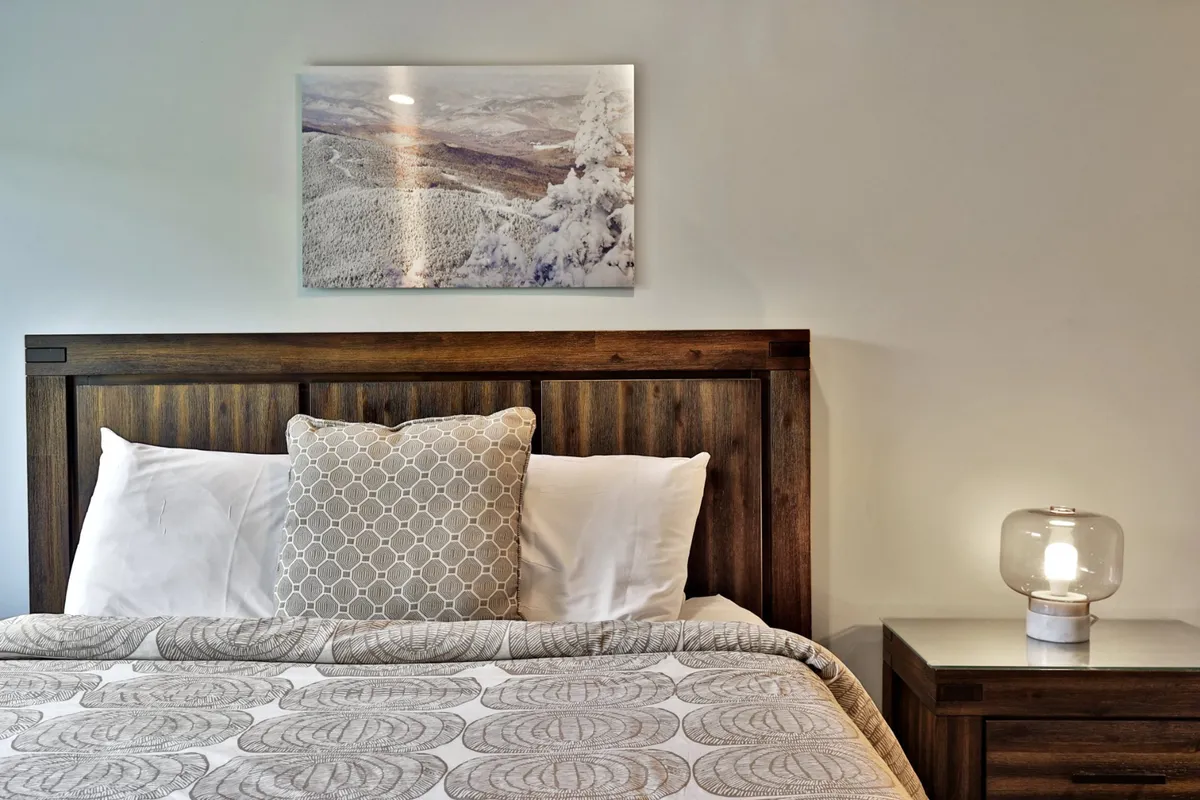 The image highlights a tranquil corner of the studio with a wooden headboard, decorative pillow, a nightstand with a lamp, and beautiful framed landscape art, suggesting a cozy and reflective space.