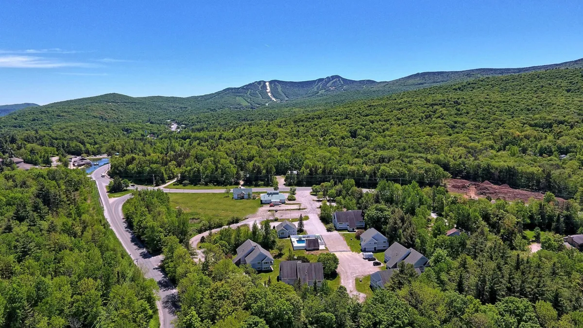 A scenic exterior shot featuring a mountain backdrop, residential buildings of the condo complex, and a roadway leading toward the slopes. The peaceful scene captures both the natural beauty of Killington and the proximity of the condo to outdoor adventure.