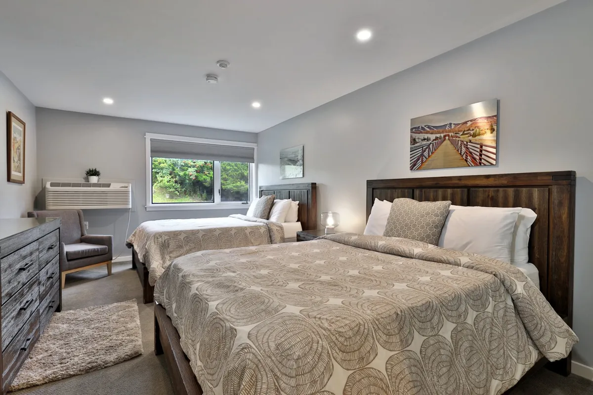 This inviting studio condo interior features two plush queen beds set against a backdrop of warm, natural light streaming through a large window. The space is decorated with tasteful artwork, a cozy atmosphere, and thoughtful amenities, creating a welcoming retreat for guests to relax and unwind after a day on the slopes.
