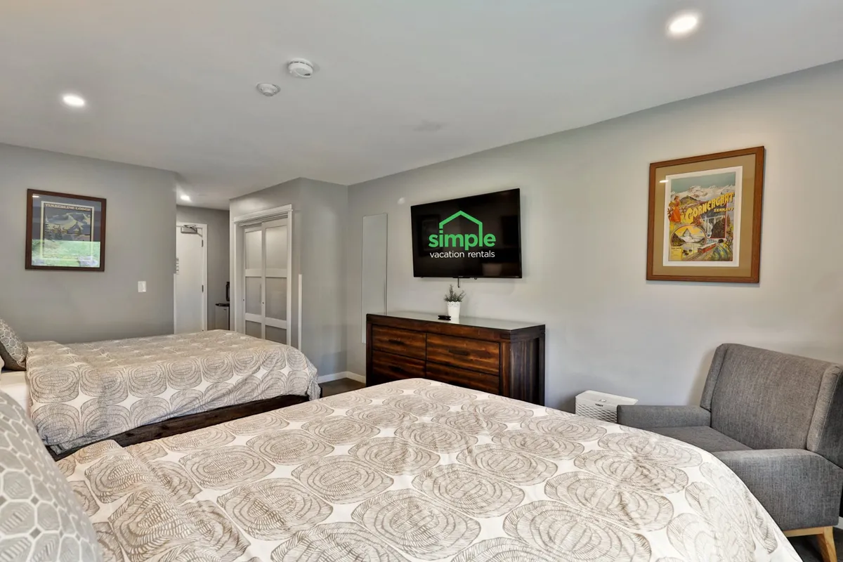 A cozy interior with two double beds, a television featuring branding, a wooden dresser, armchair, and wall artwork. The space is bright, inviting, and designed for relaxation with comfortable furnishings.