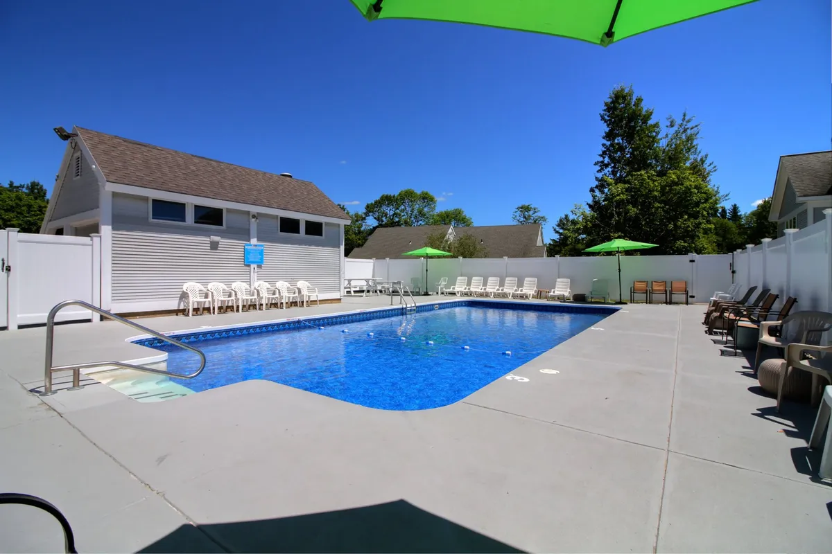 A bright, inviting outdoor swimming pool area, featuring clear blue water, a row of lounge chairs, and green umbrellas set up for shade. The setting is sunny and cheerful, suggesting a lively place for guests to swim, sunbathe, and socialize during the warmer months.