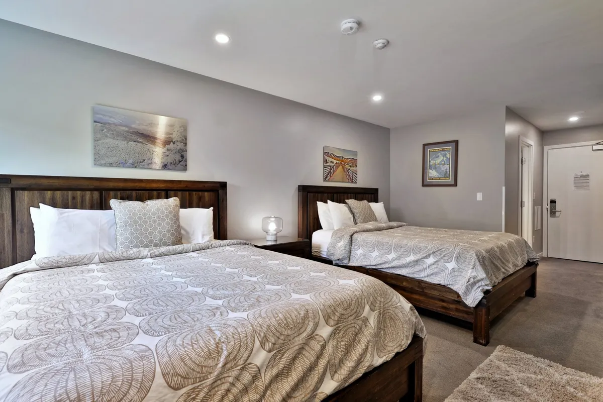 A comfortable studio space featuring two beds with plush bedding, decorative artwork on the wall, and a cozy nightstand with a lamp. The space is inviting and reflects a family-friendly environment, ideal for relaxation after a day of adventure.
