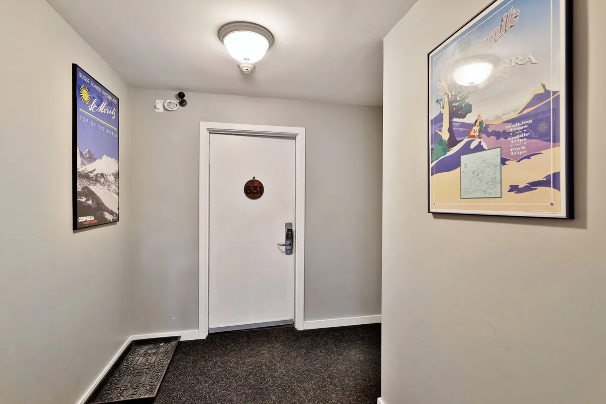 This image shows a welcoming condo entry door, marked with the number 63. The entryway is neat, with a carpet mat and framed posters, evoking a sense of arrival and transition into a private, well-managed space.