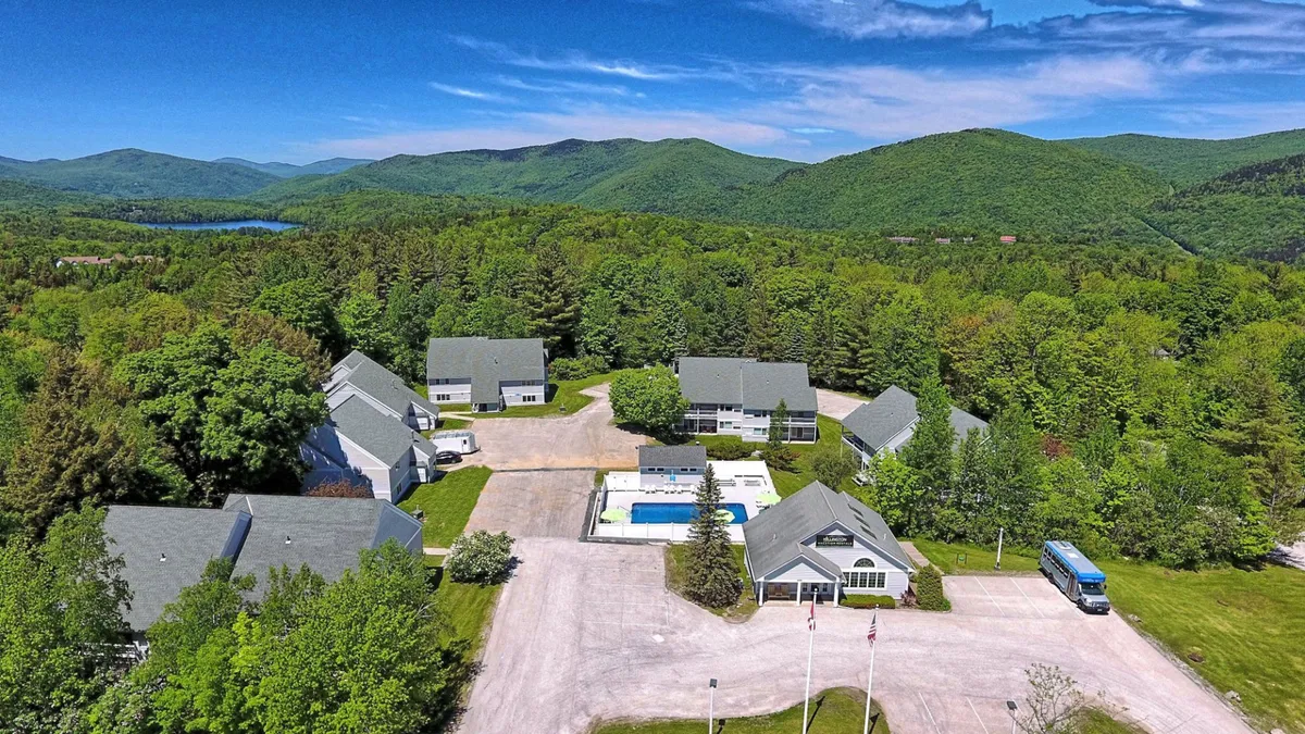 This wide, exterior shot showcases the swimming pool with a backdrop of rolling mountains, lush greenery, and the property buildings. The pool is central, surrounded by nature and offering a sense of place within the Vermont landscape.