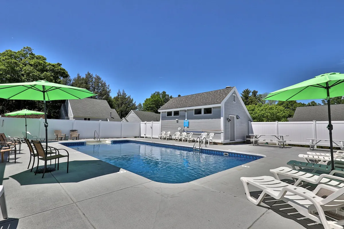 This exterior shot features the saltwater swimming pool surrounded by green umbrellas, lounge chairs, and mature trees. The pool house is visible in the background, and the setup conveys a welcoming, refreshing space ideal for relaxation and social gatherings.