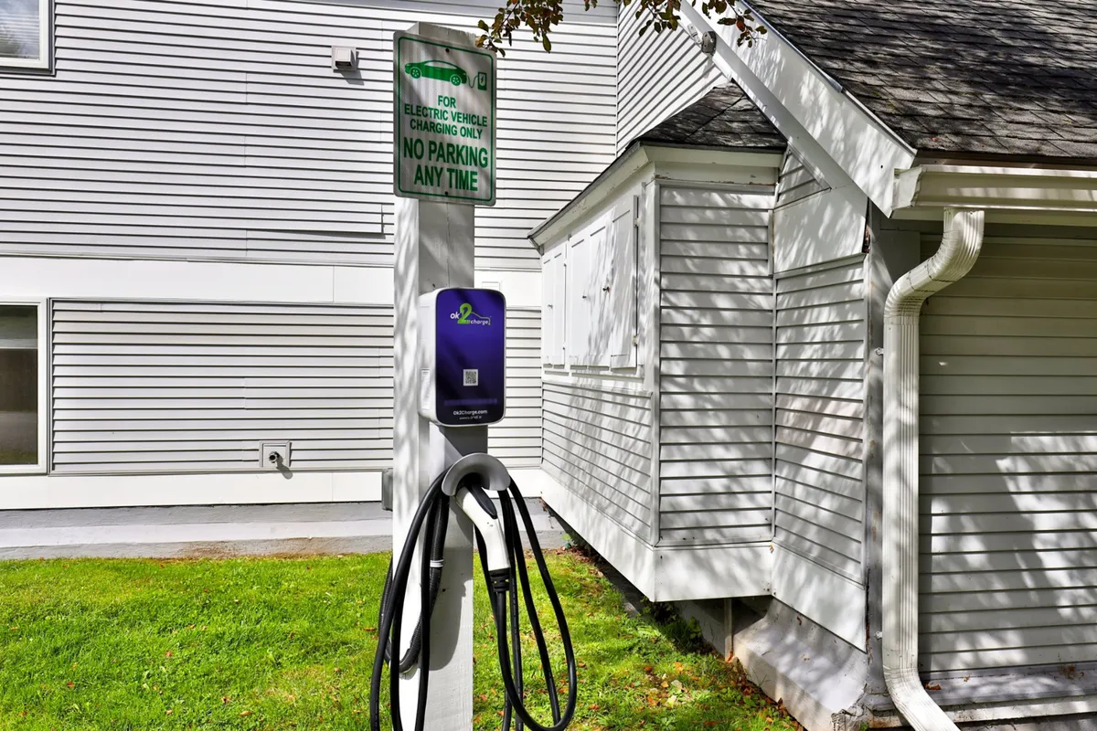 This image features the exterior of the property, showcasing an electric vehicle charging station, clear building signage, and a glimpse of the building’s entrance area. The background hints at a well-maintained setting, inviting guests to arrive or set out for the evening.